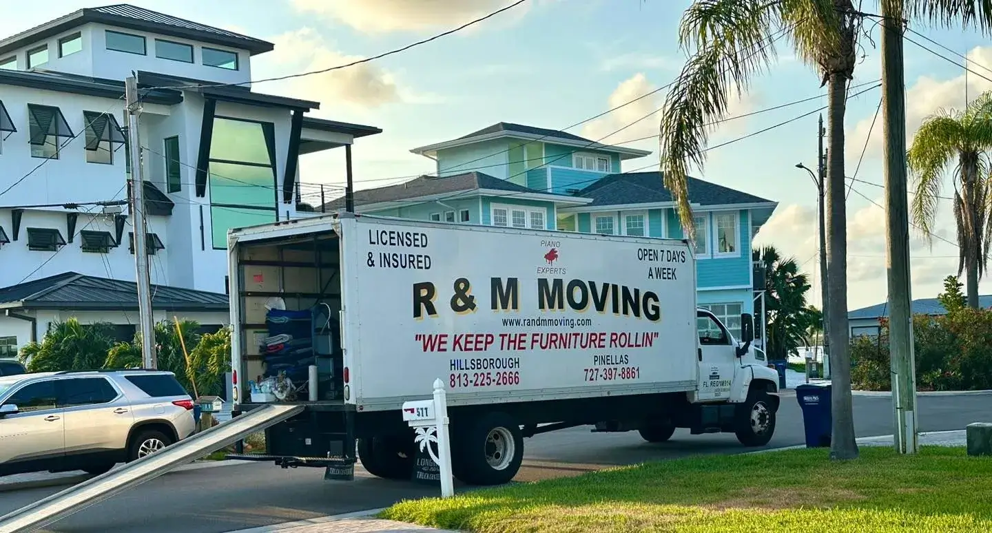 Branded R&M Moving box truck with open loading ramp parked outside a coastal Florida home.