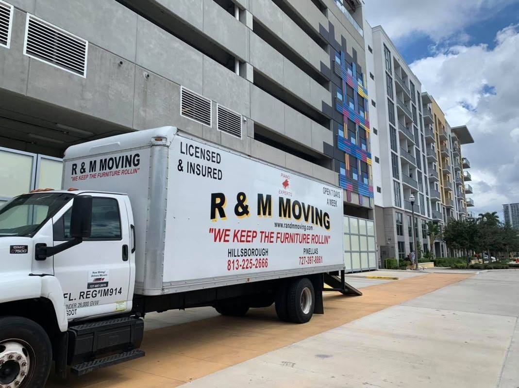 An R & M Moving truck parked outside a multi-story modern apartment building next to a concrete parking structure.