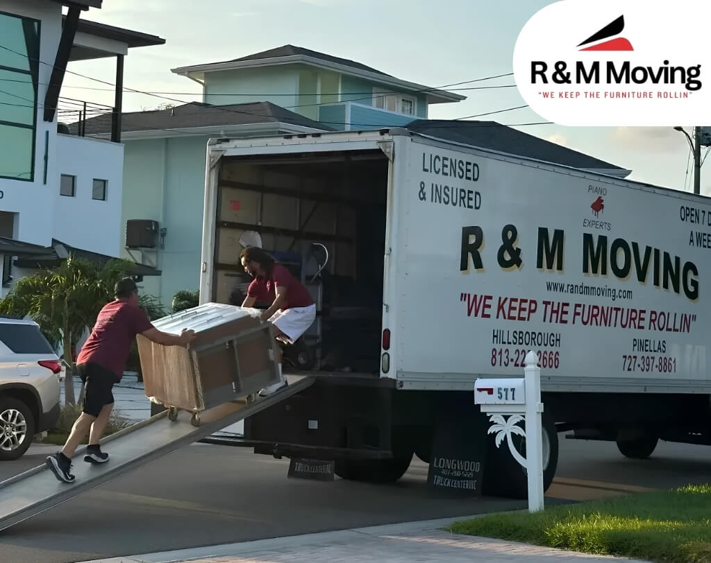 Street scene at a curb with a small truck and a black trailer, a sign reading “R&M Moving” on the side, and pedestrians and motorcycles passing by under overhead wires.