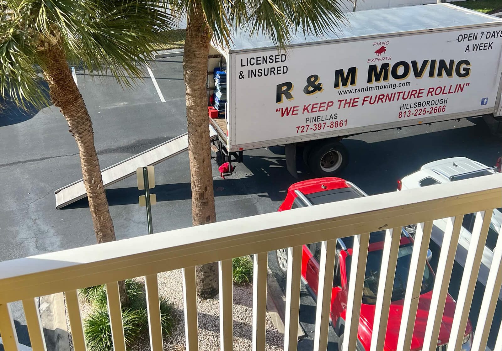 Elevated balcony view of an R & M Moving truck with ramp extended in a parking area surrounded by palm trees.