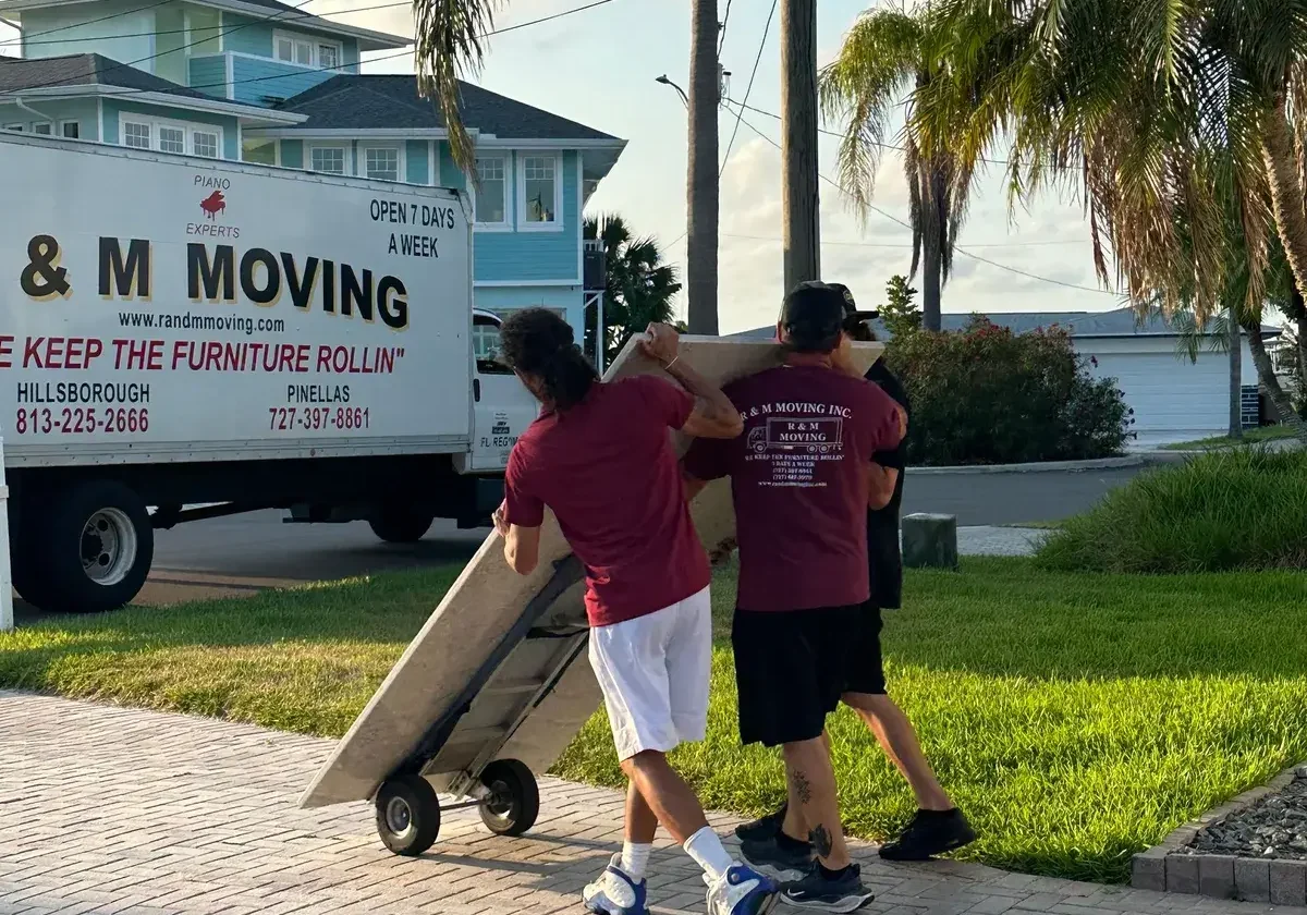 Alt Text: Three R&M Moving crew members wheeling a heavy tabletop on a hand truck along a brick driveway.