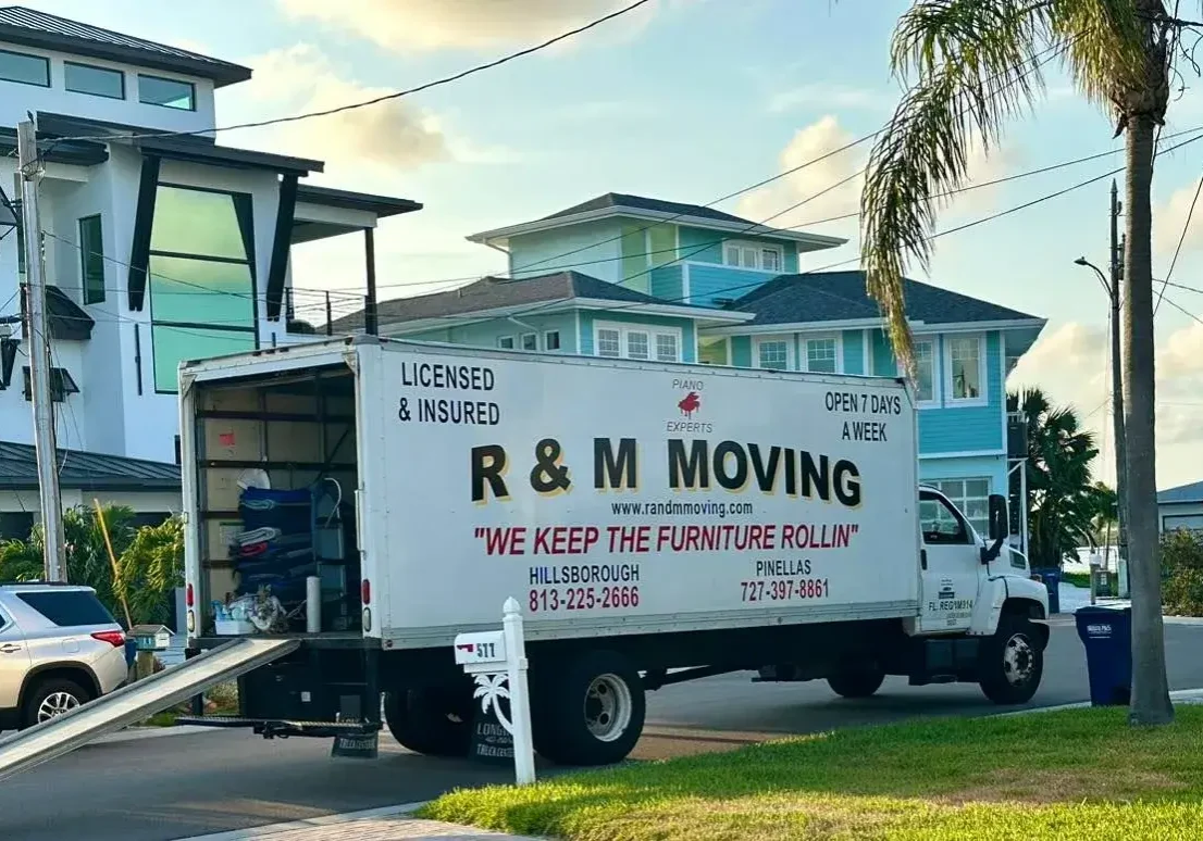 Branded R&M Moving box truck with open loading ramp parked outside a coastal Florida home.