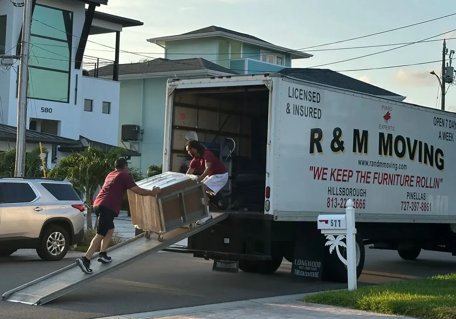 Two R&M Moving professionals loading heavy furniture up a ramp into a branded moving truck.
